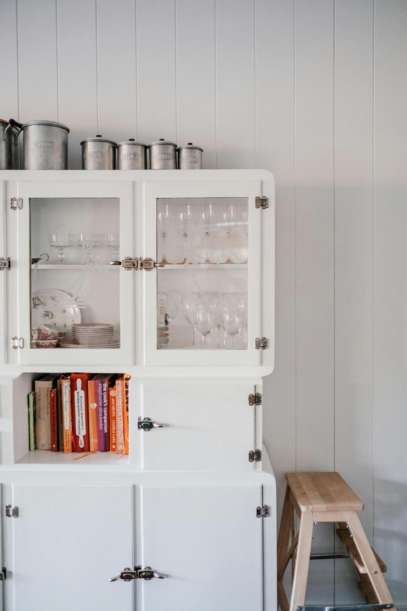 White rustic kitchen cabinet displaying glassware and cookbooks, with a wooden stool nearby.
